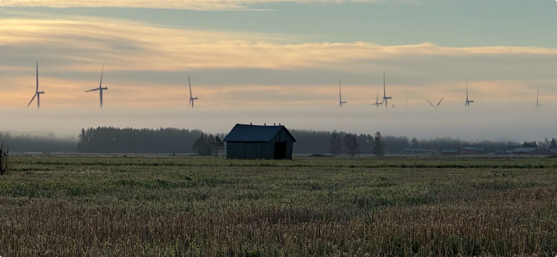 En liten lada ligger mitt på ett fält under en molnig himmel i gryningen eller skymningen, med flera vindkraftverk synliga på avstånd i ett lager av dimma.