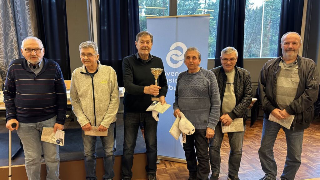 Sex äldre män står på rad inomhus, varav en håller i en pokal. De poserar för en gruppbild framför en blå banderoll med vit text och symboler. Några håller i papper, och alla är klädda i vardagskläder.