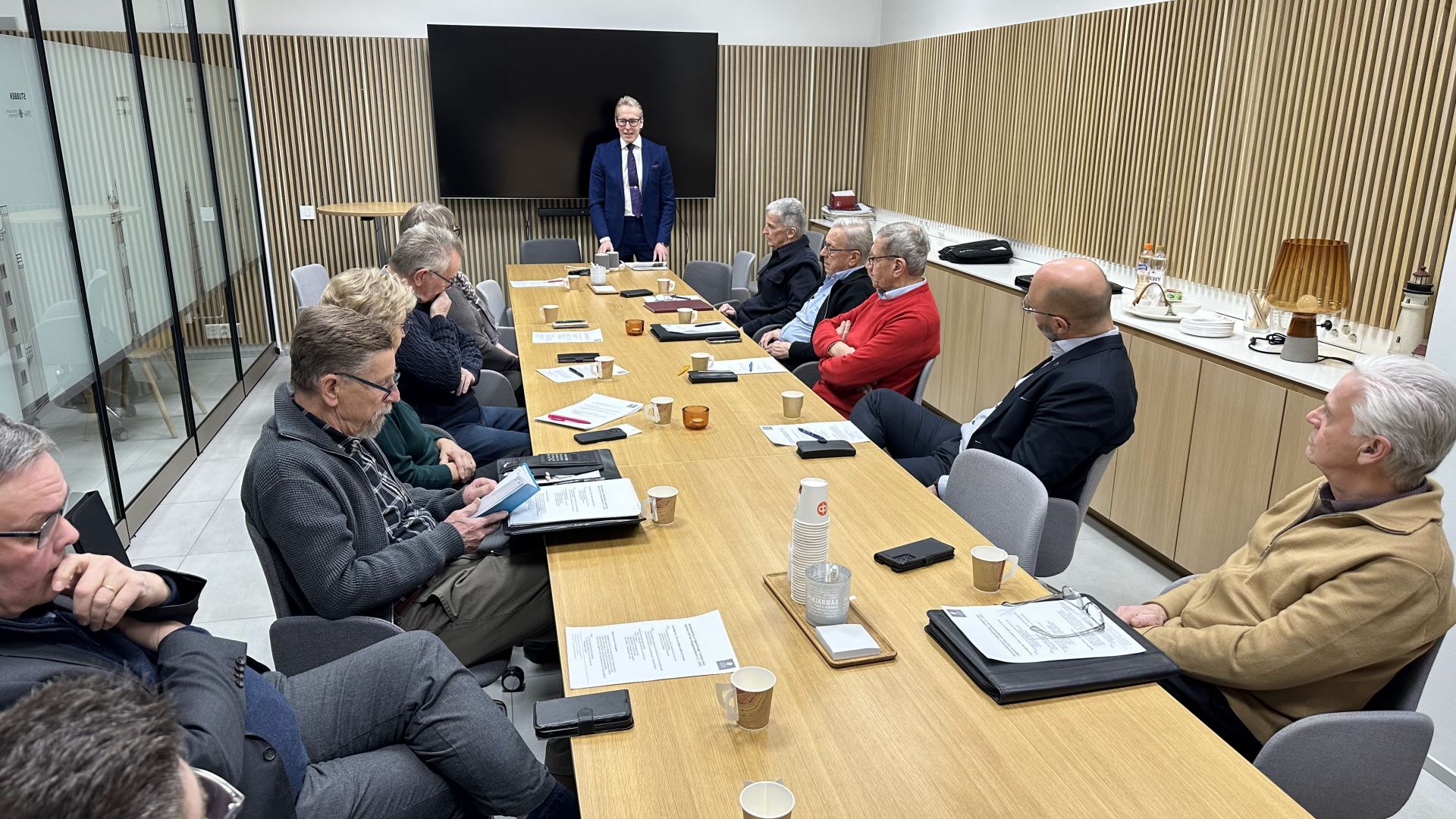 En grupp människor sitter runt ett konferensbord med dokument och kaffekoppar i ett modernt mötesrum; en person står vid bordets huvud och talar till gruppen.