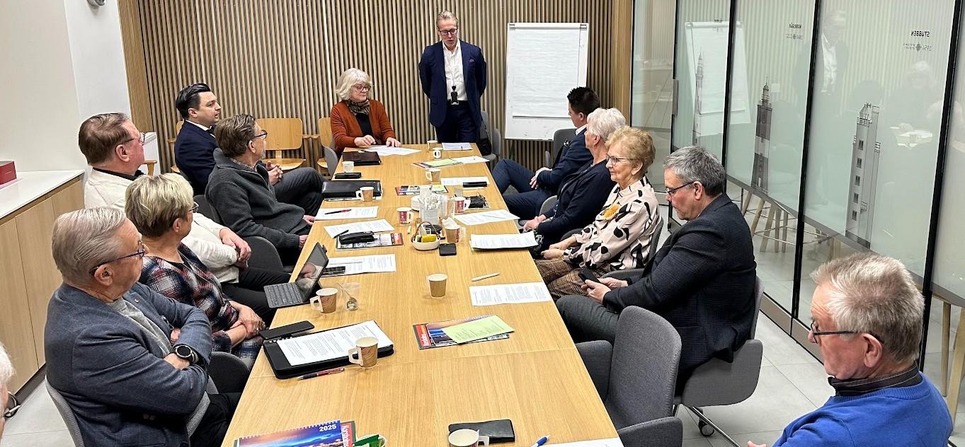 Tolv personer sitter runt ett konferensbord med papper, anteckningsblock och muggar, medan en person står vid bordsänden nära ett tomt blädderblock och talar till gruppen i ett modernt mötesrum.