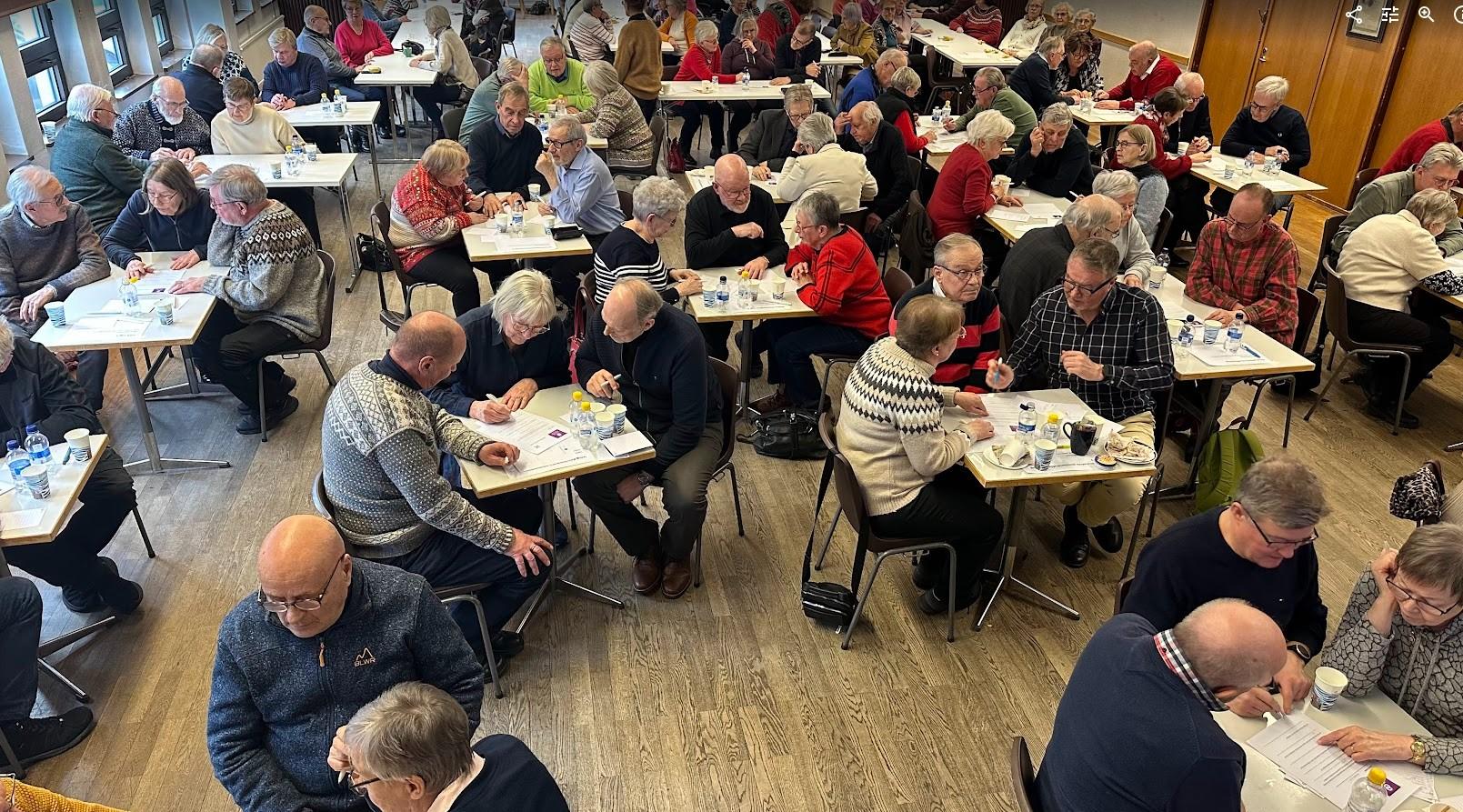 En stor grupp äldre vuxna sitter vid små bord i en sal, engagerade i samtal och aktiviteter, med papper, pennor och drycker på borden. Rummet har trägolv och fönster längs ena väggen.