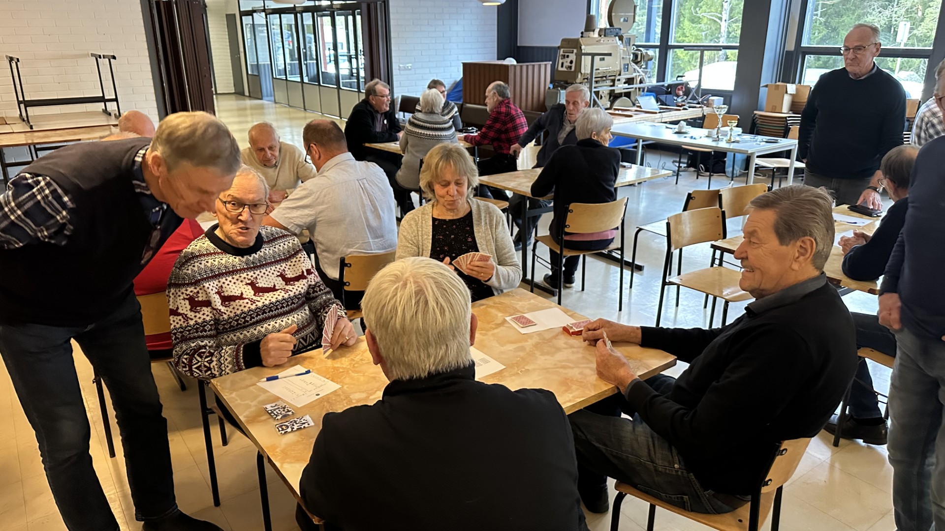 En grupp äldre vuxna sitter runt bord i en samlingslokal, spelar kort och umgås. Några personer står och tittar på. Rummet har stora fönster och en ljus, avslappnad atmosfär.