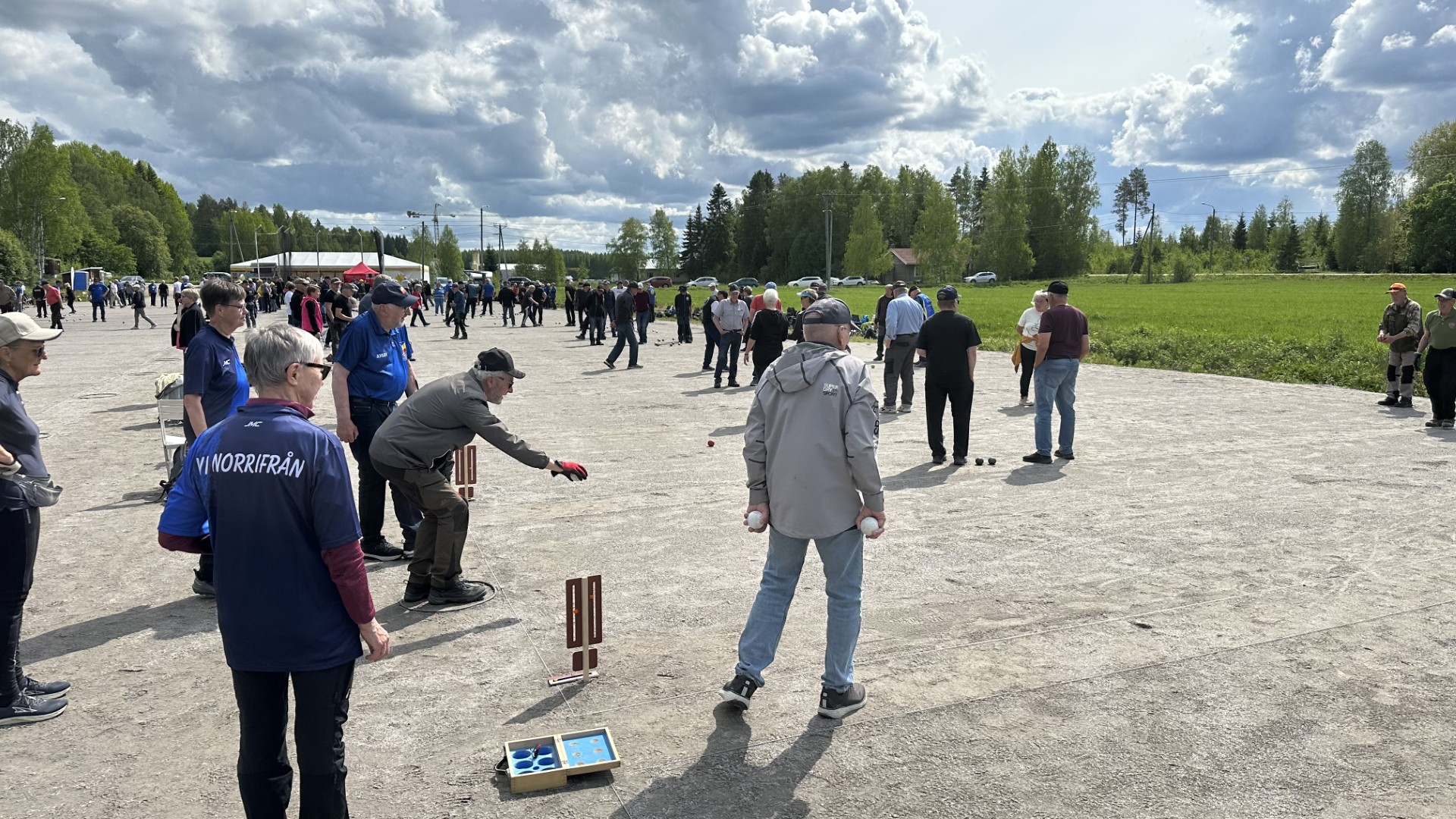 Människor samlas utomhus på en grusplan för att spela petanque. Spelarna står i grupper, vissa kastar metallkulor, medan andra tittar på. Träd och gräs kantar området under en molnig himmel.