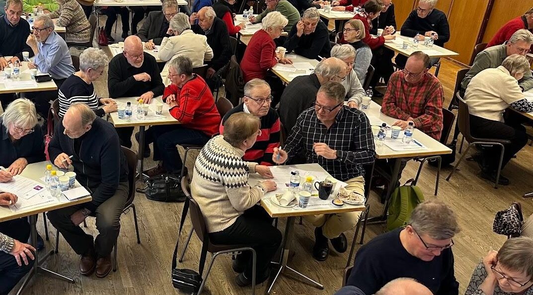 En stor grupp äldre sitter två och två vid bord i ett trångt rum och pratar och skriver på papper. Drycker, snacks och pennor finns på borden. Människorna verkar delta i en organiserad aktivitet.