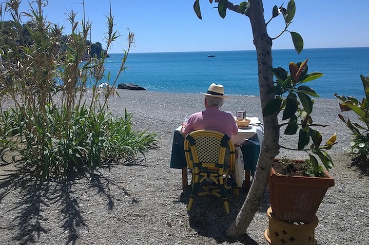 En äldre person med hatt sitter ensam vid ett bord vid stranden, vänd mot det blå havet, med växter och ett träd i närheten under en klar himmel.