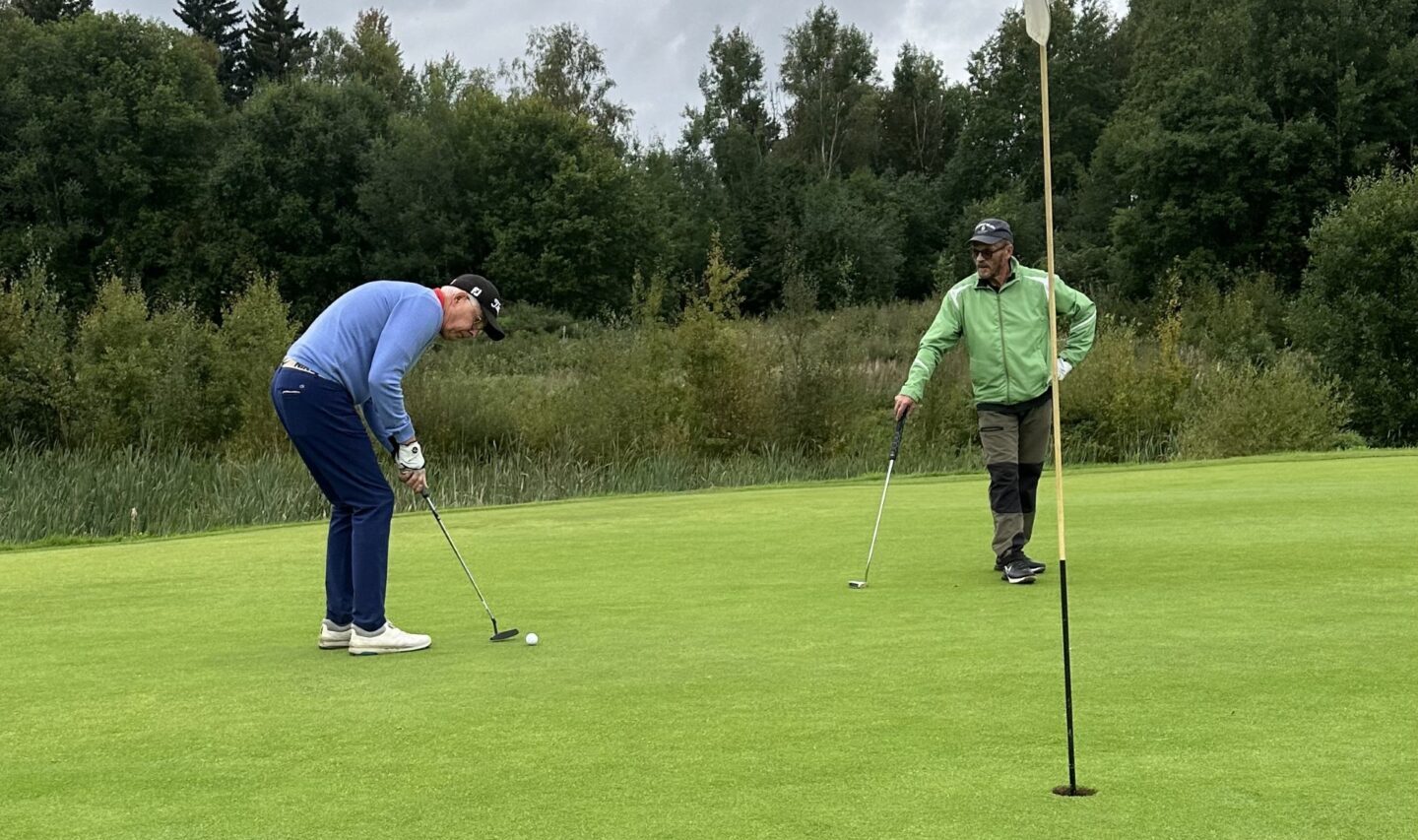 Två personer befinner sig på en golfgreen; den ena puttar medan den andra står bredvid och tittar på. En flagga är i hålet och i bakgrunden syns träd och en molnig himmel.