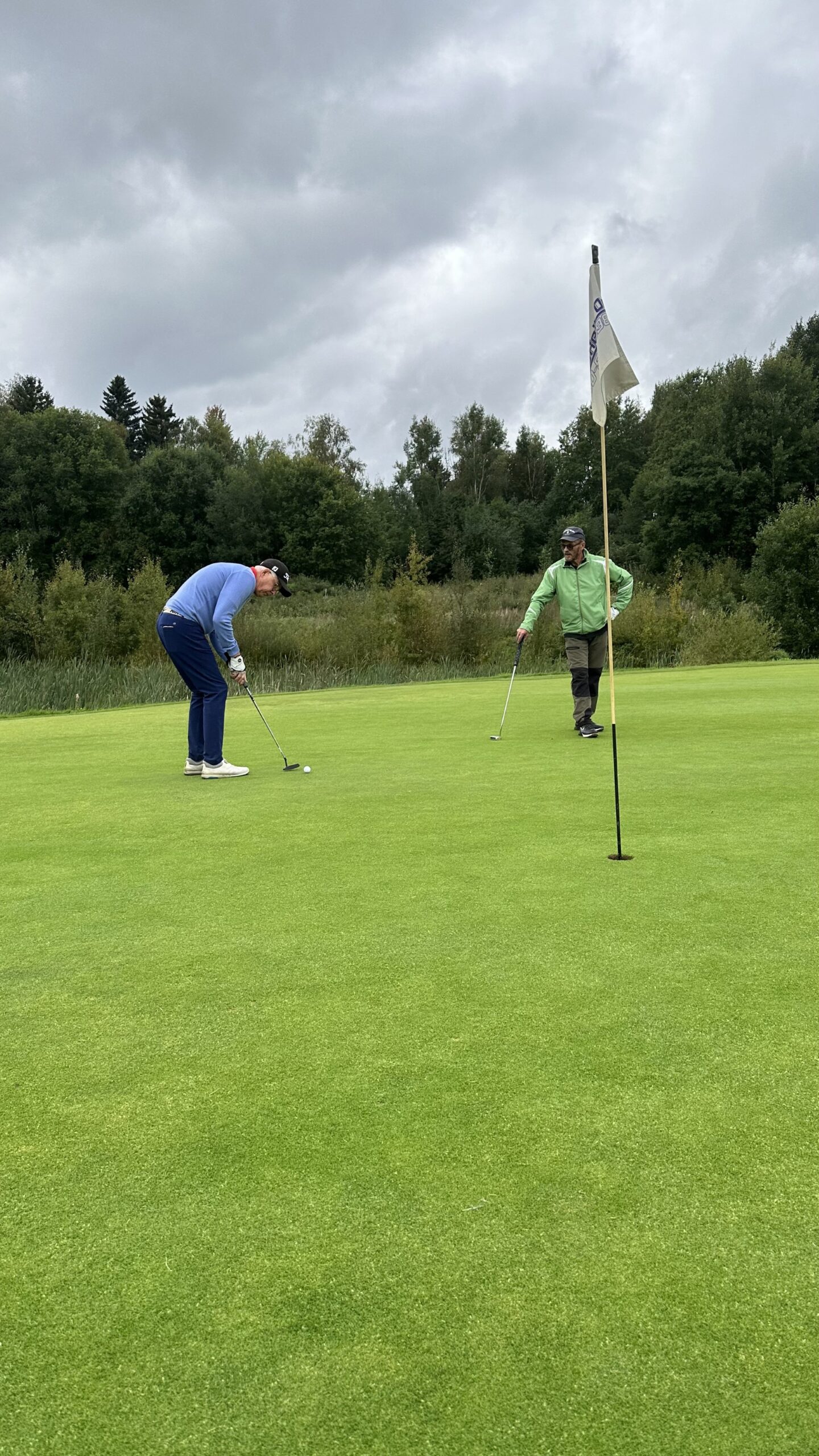 Två personer befinner sig på en golfgreen; den ena puttar medan den andra står bredvid och tittar på. En flagga är i hålet och i bakgrunden syns träd och en molnig himmel.