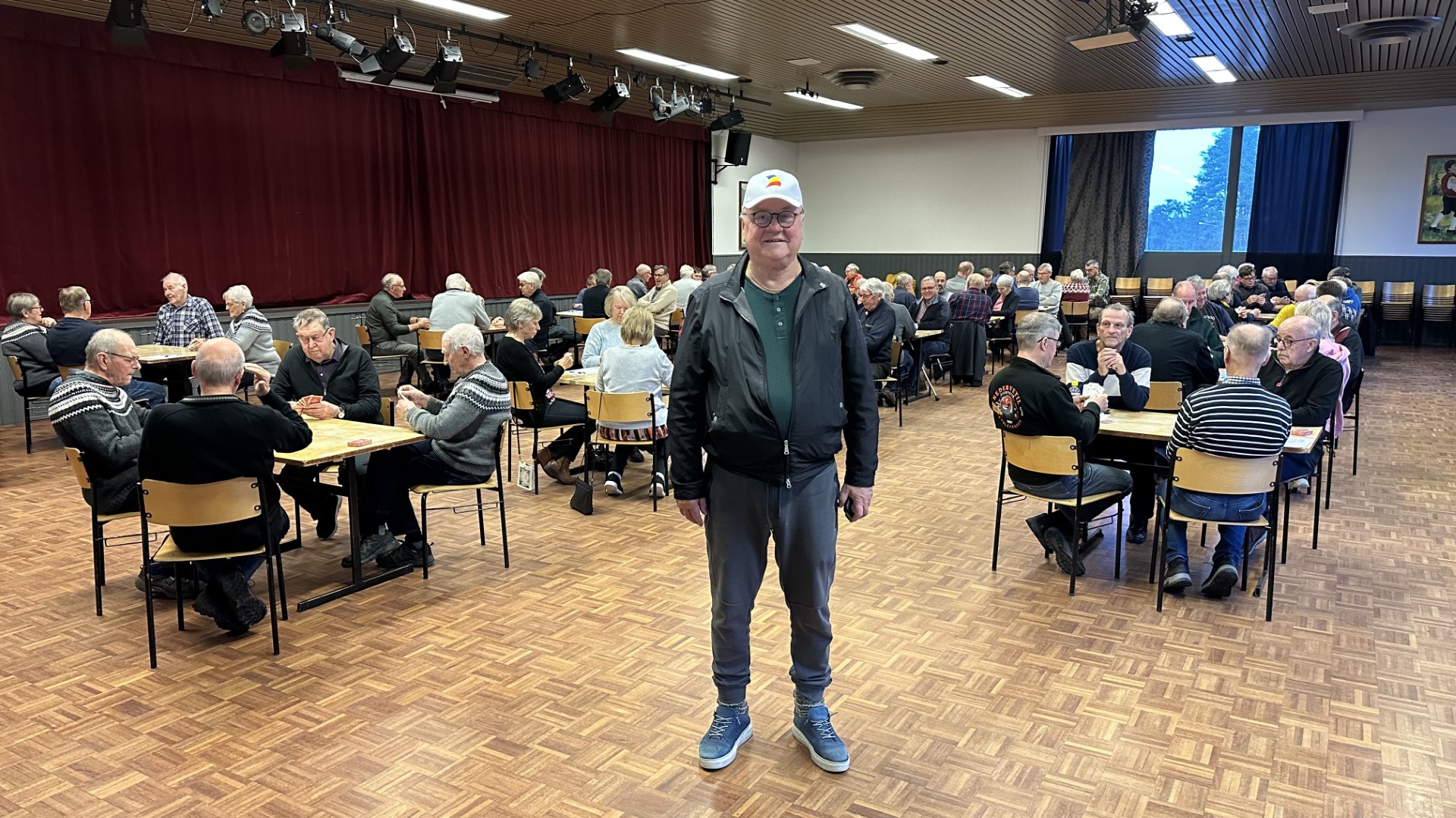 En man i kavaj, grön skjorta och vit keps står i mitten av ett stort rum. Runt borden sitter grupper av människor, engagerade i samtal eller aktiviteter. Rummet har trägolv och röda gardiner på baksidan.