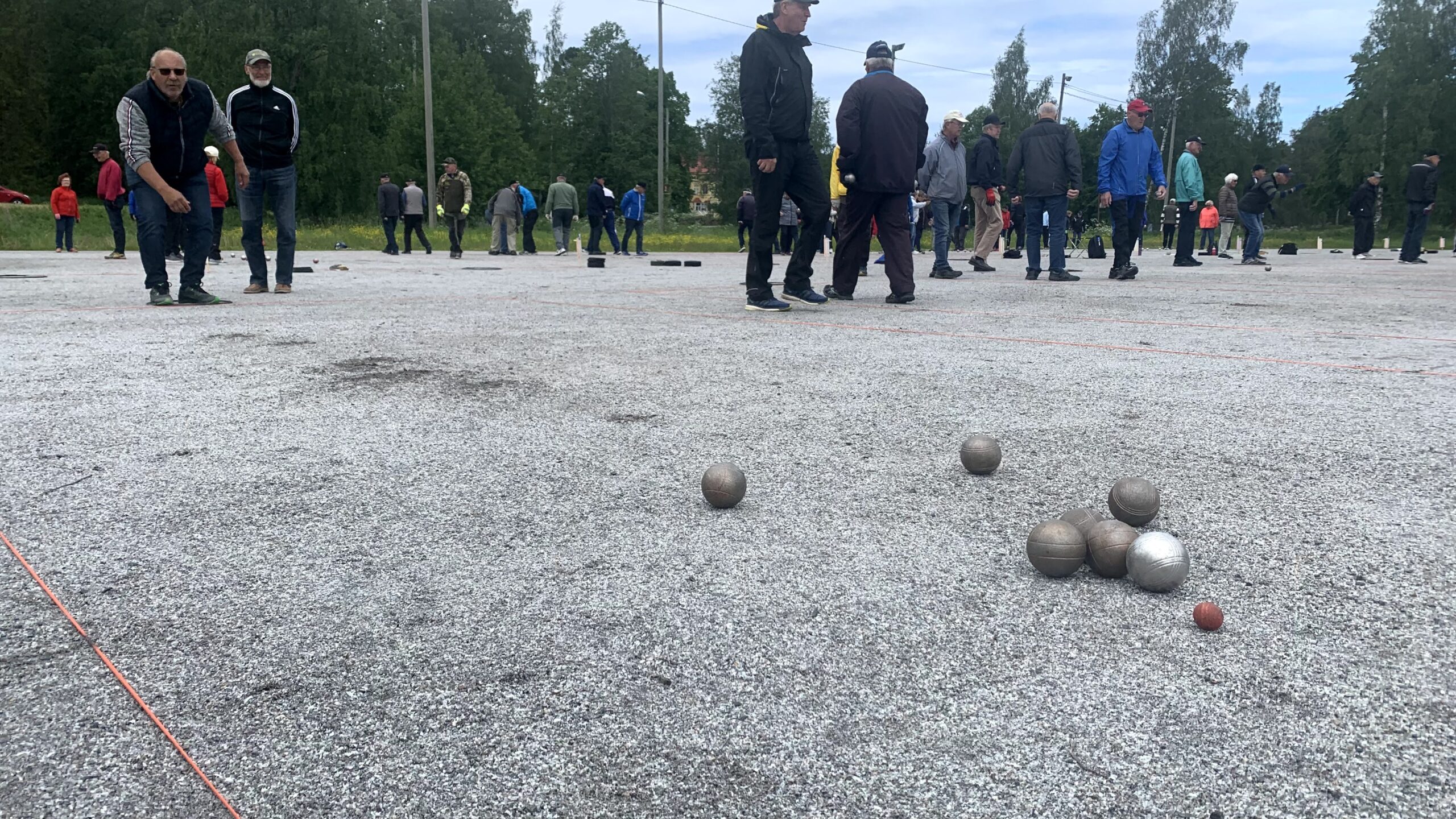 En grupp människor spelar petanque på en grusplan utomhus. Flera metallkulor och en liten målkula syns på marken i förgrunden. Träd och gräs syns i bakgrunden.