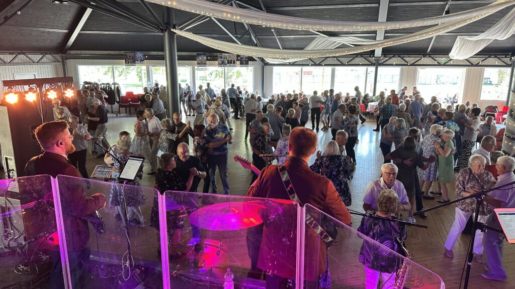 Ett band uppträder på scenen i en stor sal med en publik bestående av främst äldre vuxna som dansar och umgås. Bilden är tagen bakom bandet och visar instrument, lampor och människor som njuter av evenemanget.
