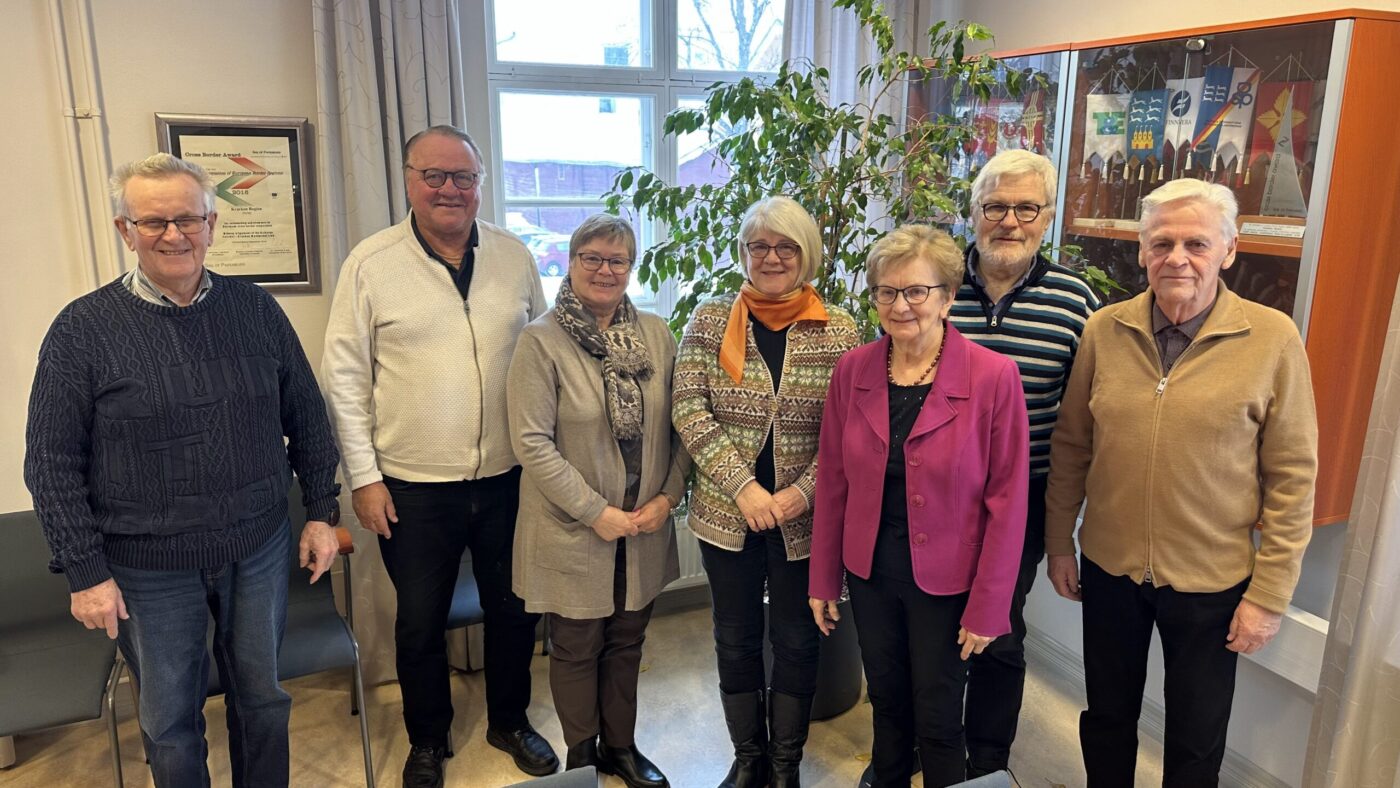 Sju vuxna står på rad inomhus och poserar för ett gruppfoto. Bakom dem finns ett fönster, en grön växt, inramade bilder på väggen och en vitrin med olika föremål. De flesta ler och har vinterkläder på sig.