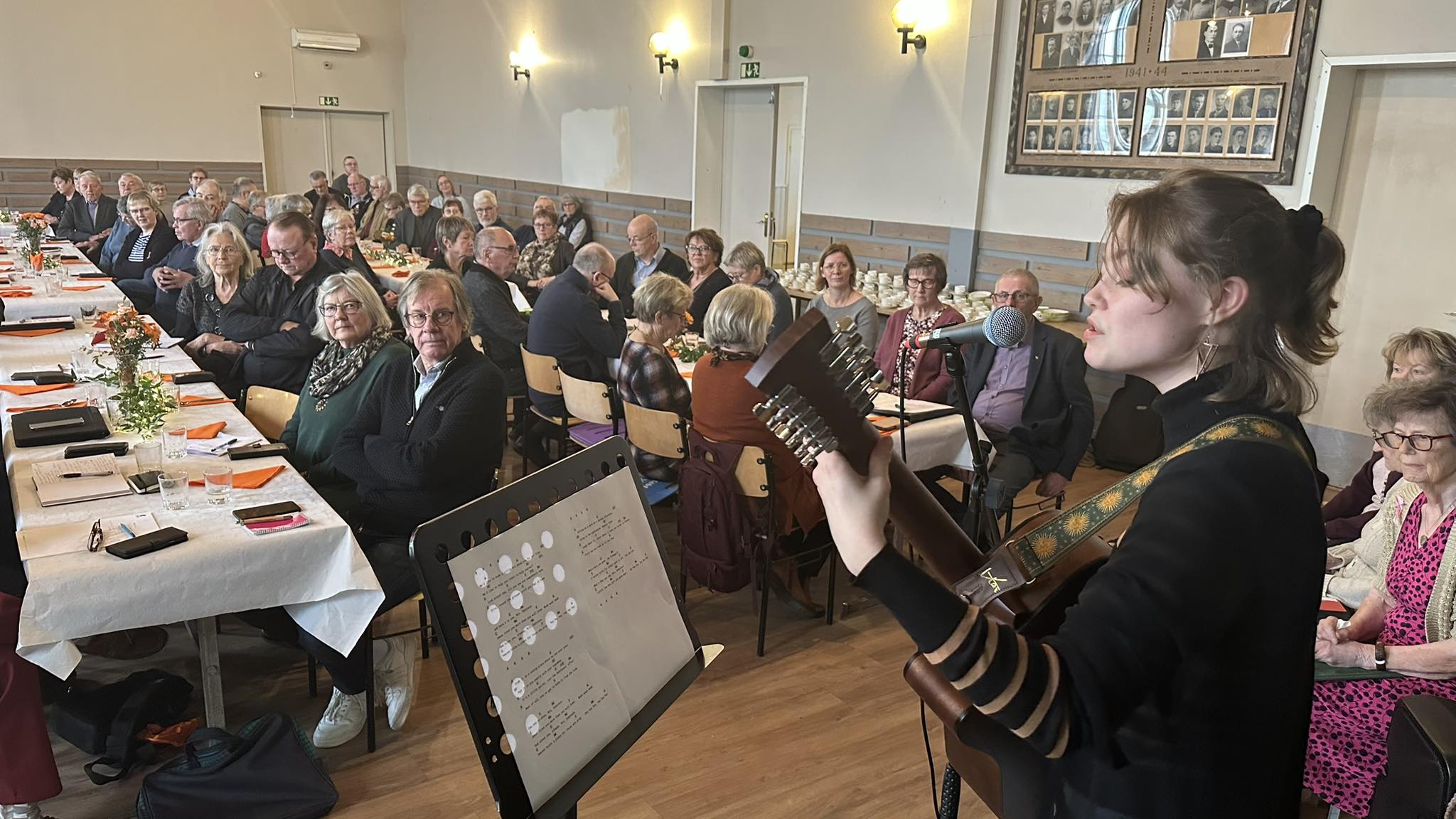 En ung kvinna spelar gitarr och sjunger i en mikrofon inför en sittande publik vid långbord i ett väl upplyst rum med inramade foton på väggen och noter på ett stativ.