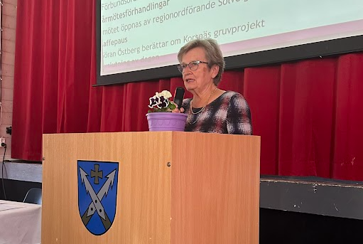En äldre kvinna står på ett podium med ett vapenemblem och talar framför en röd gardin. En lila krukväxt står på podiet. Bakom henne syns en projicerad presentation med svensk text.
