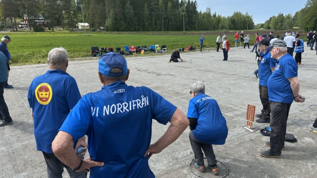 En grupp människor, främst äldre vuxna, klädda i likadana blå tröjor med text på ryggen, står och tittar på en utomhusmatch på en betongyta omgiven av en gräsplan och träd.