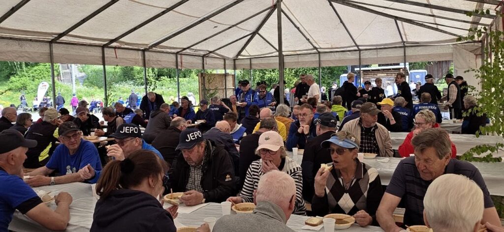 En stor grupp människor, främst äldre, sitter och äter vid långa bord under ett tält. Många har på sig hattar och jackor, och stämningen tyder på ett gemenskapsarrangemang eller en sammankomst. Utanför tältet syns träd och grönska.