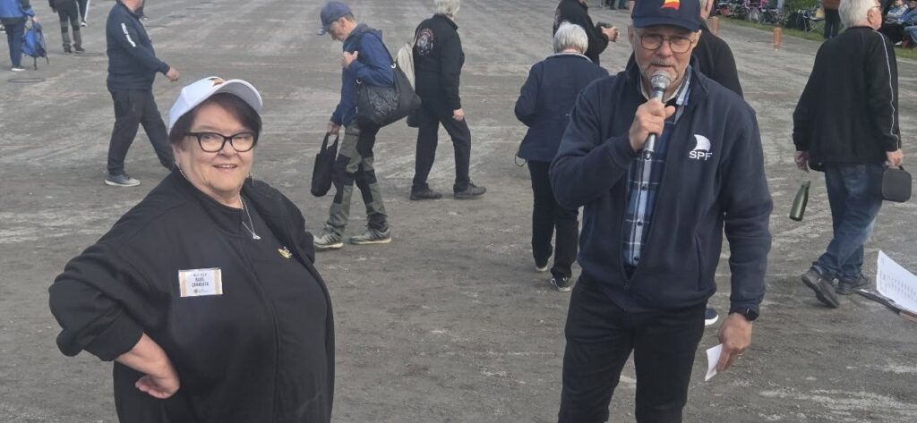 En kvinna med glasögon och en vit keps står bredvid en man som håller i en mikrofon och några papper. I bakgrunden går flera personer på en stor, öppen, asfalterad yta.