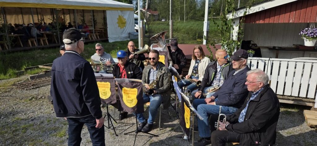 En grupp musiker med bleckblåsinstrument sitter utomhus i en halvcirkel, vända mot en dirigent. De befinner sig intill en vit och röd träbyggnad, med ett tält och sittande människor i bakgrunden.