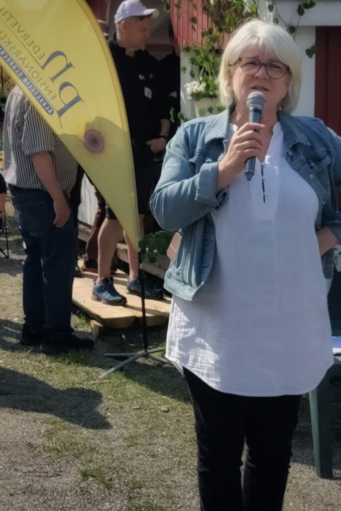 En kvinna med vitt hår och glasögon håller i en mikrofon och talar utomhus. Hon har på sig en vit skjorta och en jeansjacka. I bakgrunden står flera personer nära en gul flagga och en träbyggnad.