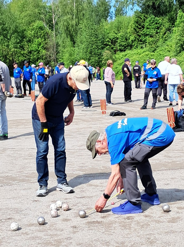 Två äldre män spelar pétanque utomhus på en asfalterad yta. Den ene mannen står upprätt medan den andre böjer sig ner bredvid flera metallbollar som ligger på marken. I bakgrunden syns andra spelare och träd.