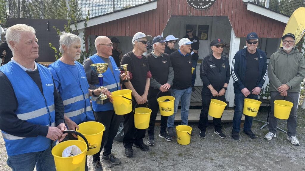 En grupp på elva män står på rad utomhus; de flesta håller i gula hinkar. Två män håller i guldtroféer. Flera av männen bär blå västar, medan andra är klädda i svarta jackor och kepsar. I bakgrunden syns en röd träbyggnad.