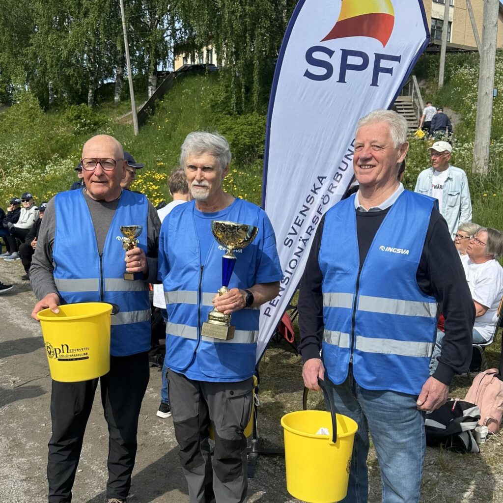 Tre äldre män i blå västar står utomhus med gula hinkar och pokaler i händerna. Bakom dem syns en flagga med texten SPF och en grupp sittande personer. I bakgrunden syns träd och en byggnad.