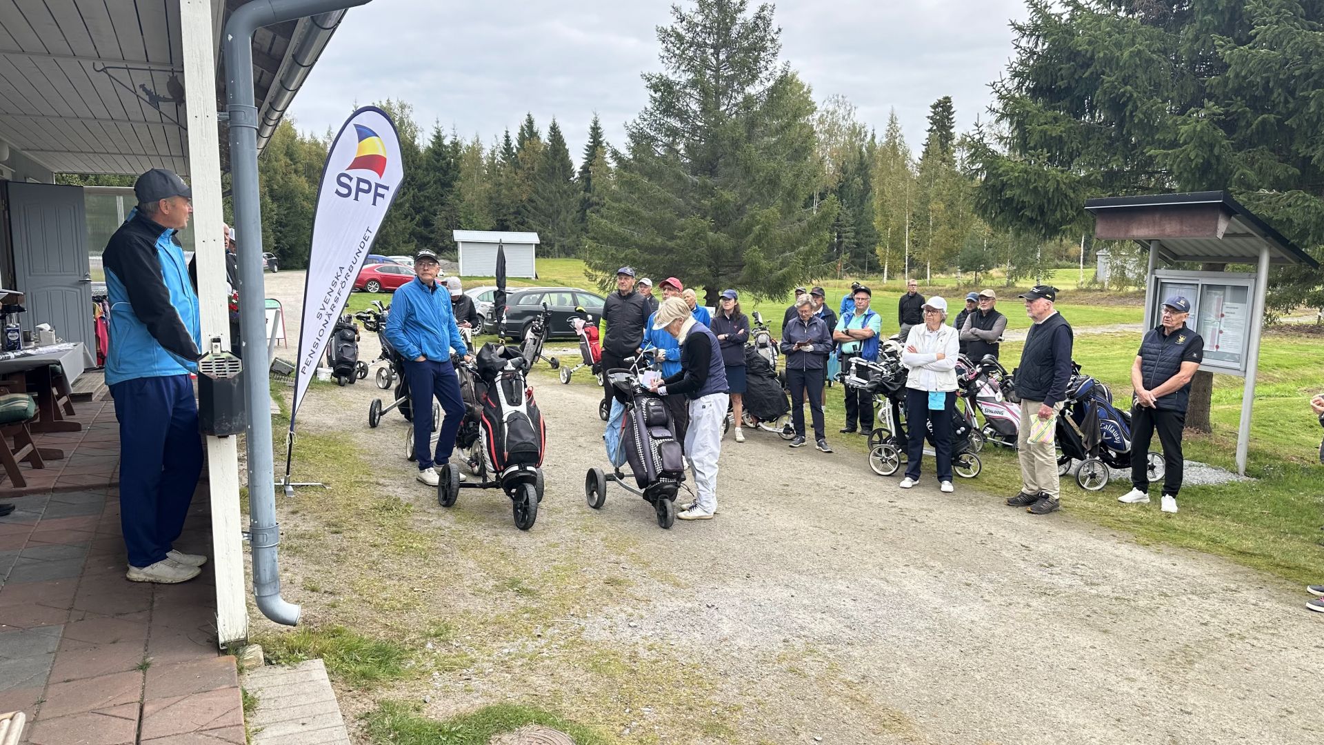 En grupp människor med golfväskor och golfvagnar står utanför nära en byggnad och en SPF-flagga och förbereder sig inför ett golftävling. Vissa lyssnar på en talare medan andra pratar med varandra. I bakgrunden syns träd och bilar.
