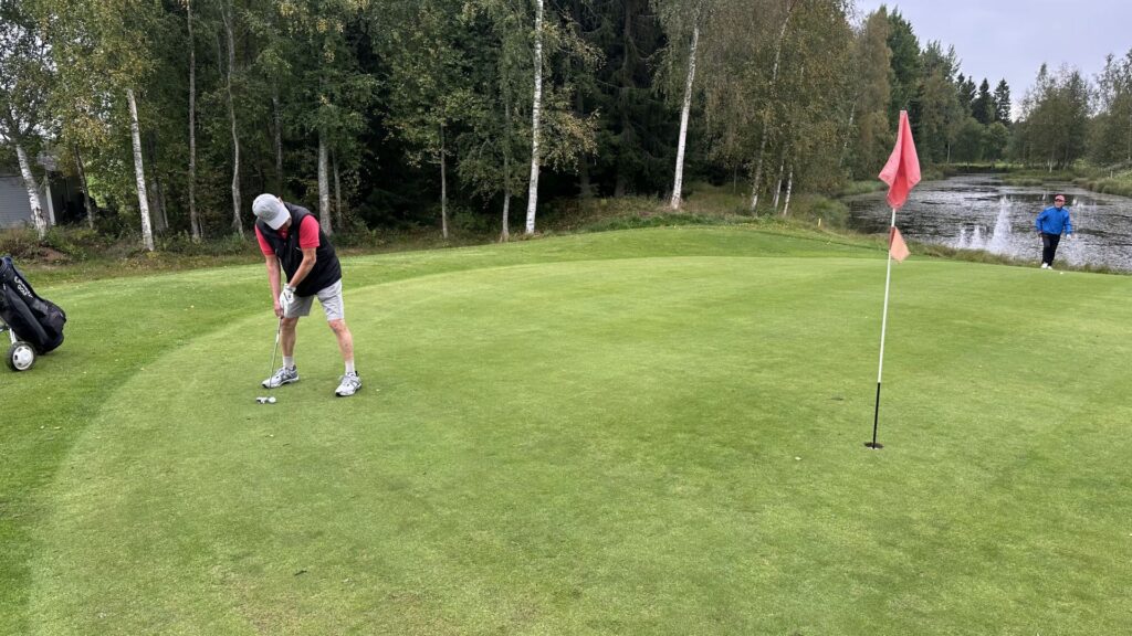 En person puttar en golfboll på en green nära ett hål med en röd flagga, medan en annan person går i bakgrunden. Bakom greenen syns träd och en vattenspegel.