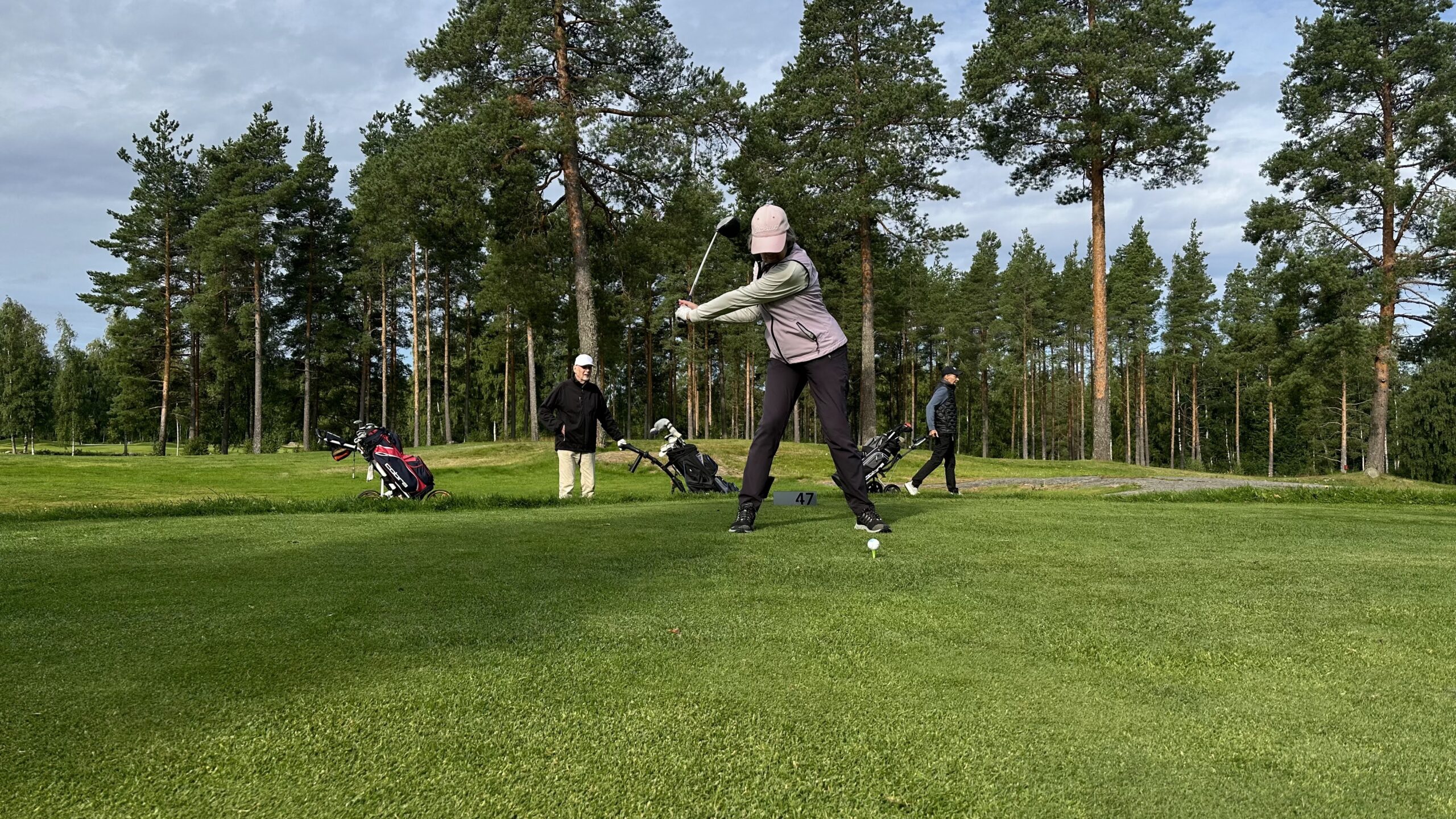 En person gör sig redo att svinga en golfklubba på en golfbana, med tre andra personer och flera golfväskor synliga i bakgrunden, omgiven av träd under en delvis molnig himmel.