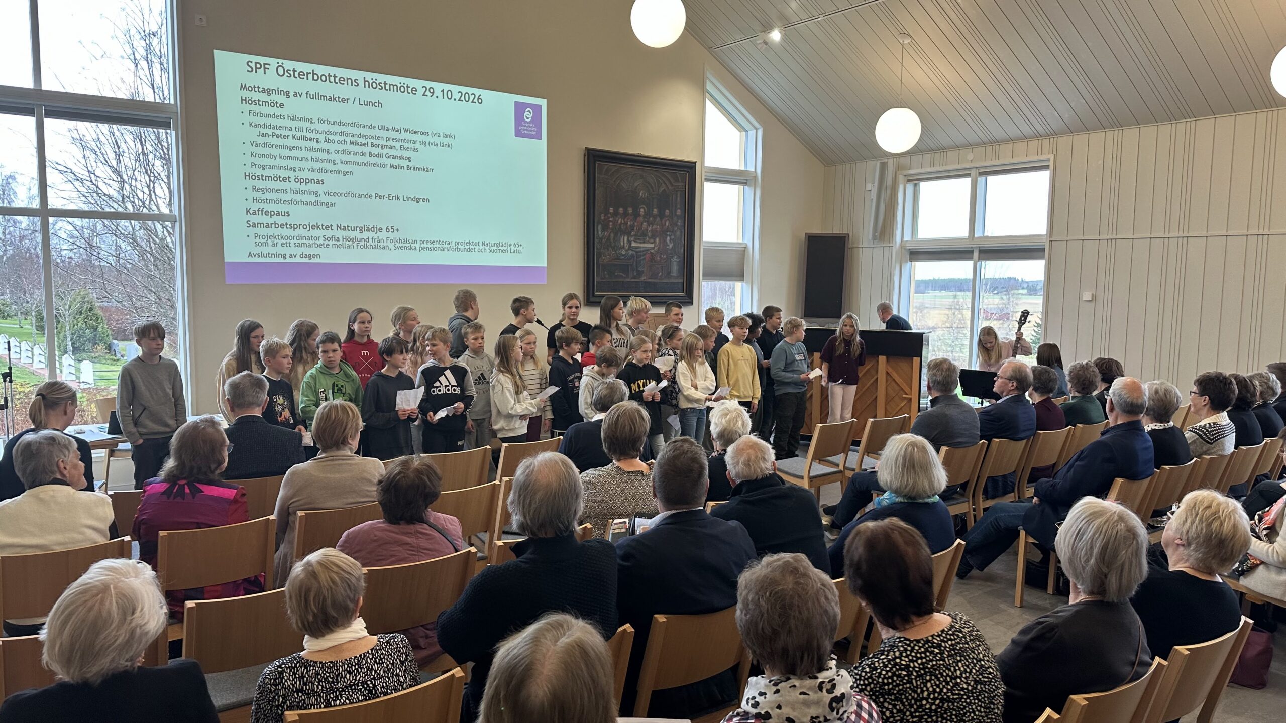 En grupp barn står framför en publik i en sal, medan en projektorskärm visar en mötesdagordning på svenska.