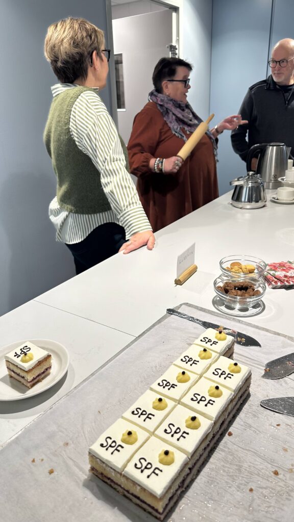 En plåtkaka skuren i små fyrkantiga bitar, toppad med vit glasyr, en gul prick och bokstäverna SPF, med en portion på en tallrik intill och tre personer som står i bakgrunden.